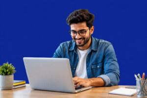 Young man working on a laptop at a modern desk with a dark blue background (#162B8D), smiling while typing in a clean professional workspace.
